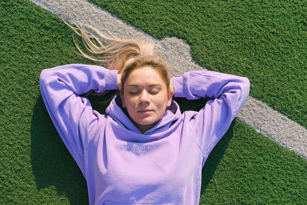 Woman in purple hoodie relaxing on green sports field under sunlight.